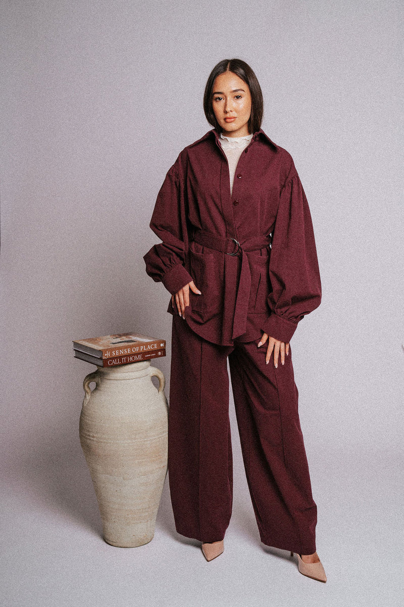 Woman in maroon belted suit stands by ceramic vase and books in minimalist studio