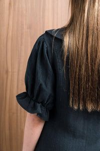 Back view of a woman in a black puff sleeve dress with straight brown hair, wood background