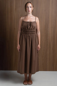 Woman in brown spaghetti strap midi dress with ruched waist, standing against wood wall