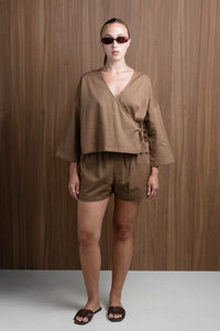 Woman in brown linen wrap top and shorts with sunglasses, standing by wood wall