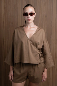 Woman in brown wrap top and shorts, tortoiseshell sunglasses, wood panel background