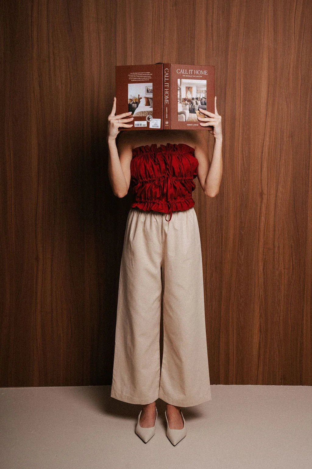 Woman in red ruched top and beige wide-leg pants holding an open book in front of wooden wall