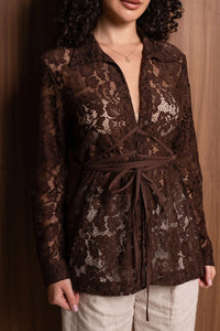 Woman in brown lace wrap blouse and cream pants, standing against wood background