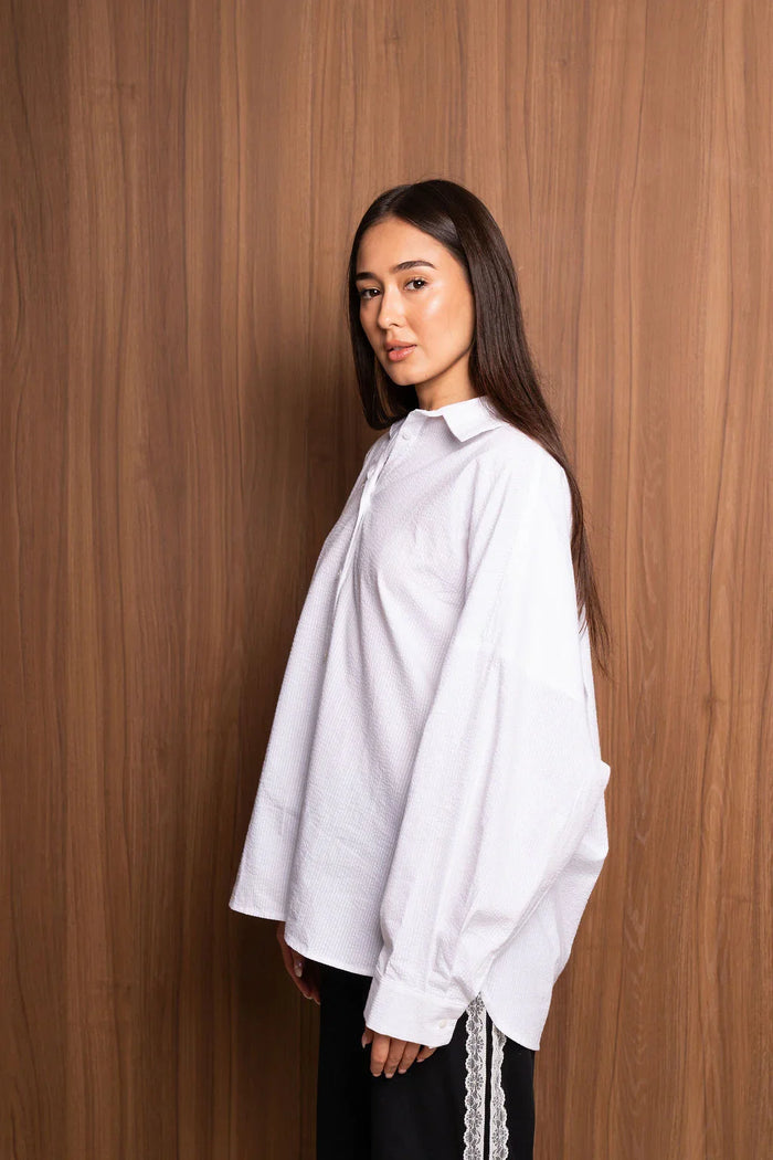 Woman in oversized white textured shirt with long sleeves, posing by wood panel wall