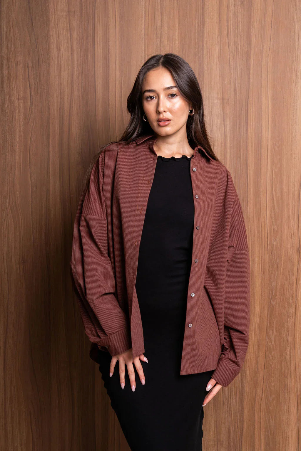 Woman in brown oversized corduroy shirt and black dress posing against wood wall