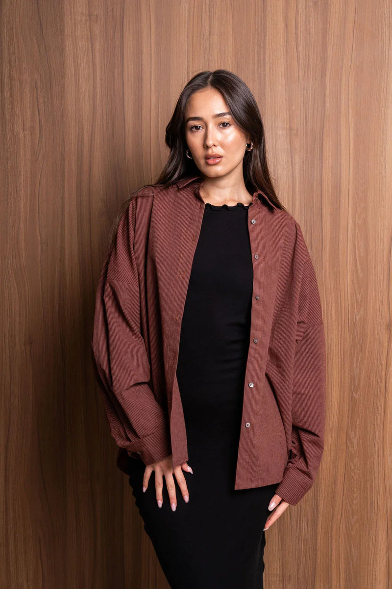 Woman in brown oversized corduroy shirt and black dress posing against wood wall