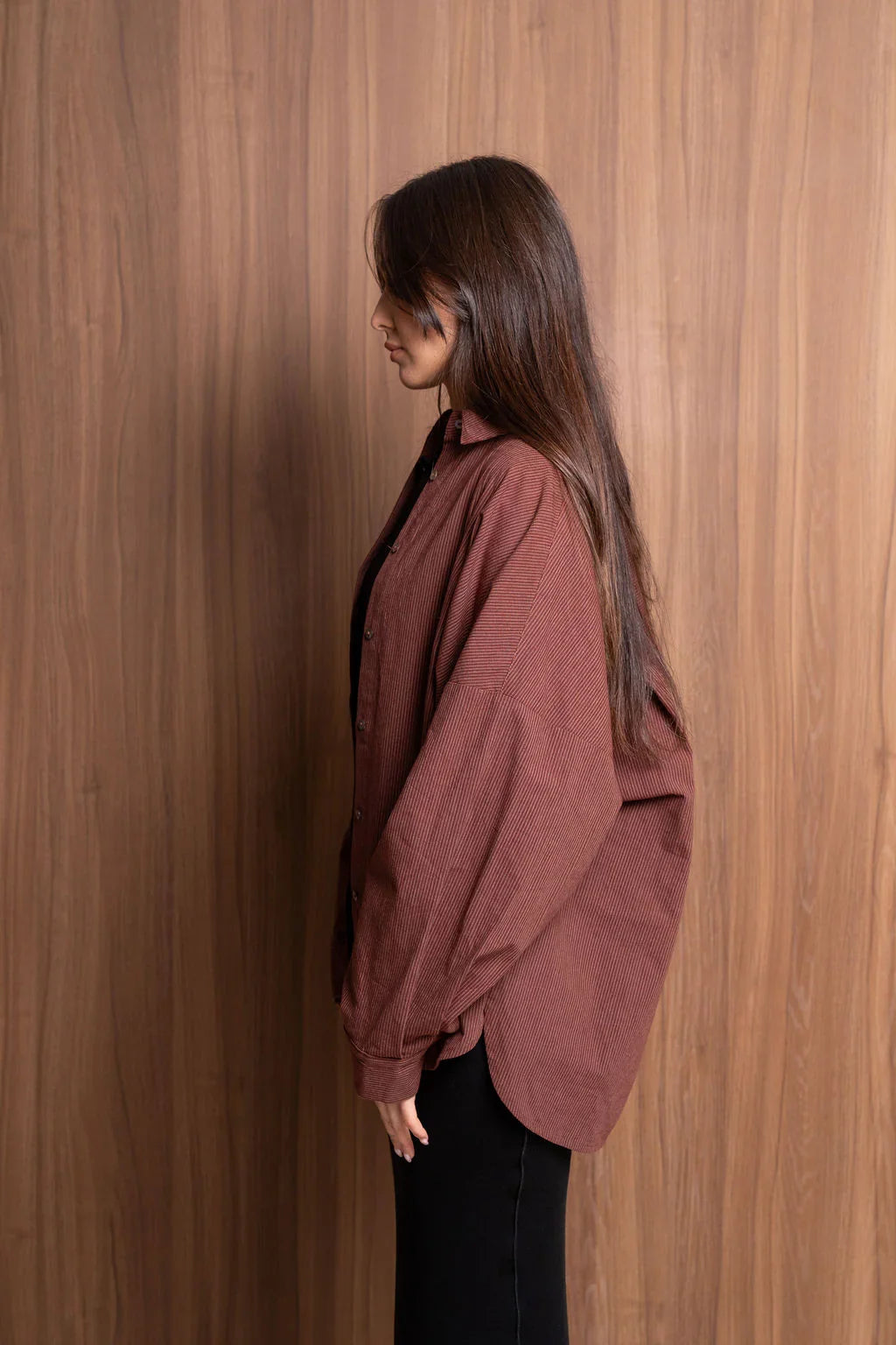 Woman in loose brown corduroy shirt and black pants standing against wood wall