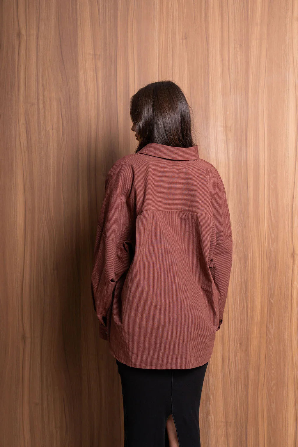 Woman in brown oversized shirt and black skirt standing against wood panel wall