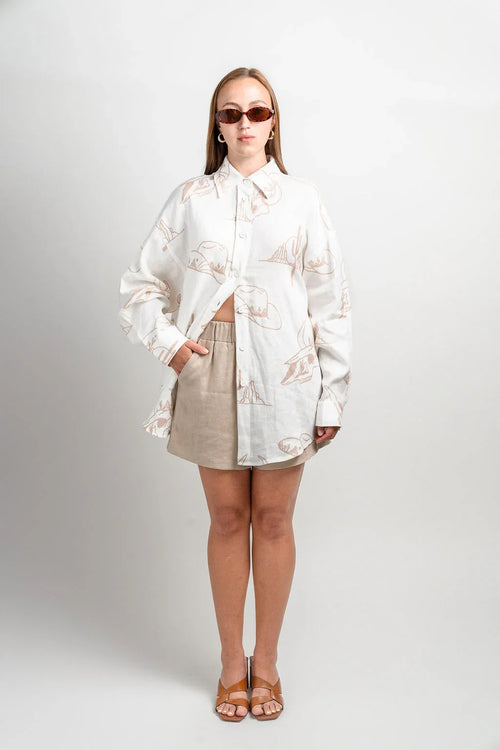 Woman wearing beige shorts and a white button-up shirt with desert print, sunglasses, sandals