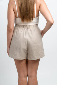 Back view of woman wearing beige linen shorts and cream tank top against white background