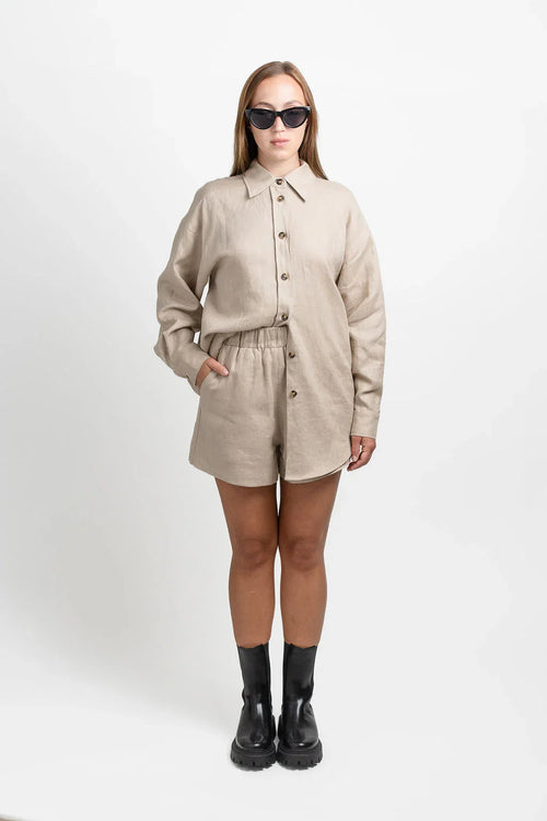 Woman in beige linen shirt and shorts set, sunglasses, and black boots on white background