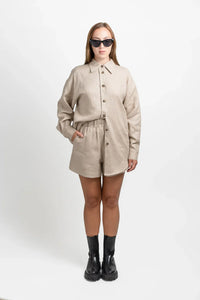 Woman in beige linen shirt and shorts with black boots and sunglasses, studio background