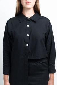 Woman wearing a black button-up shirt with white buttons, long sleeves, plain background