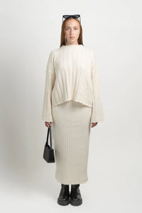 Woman in ivory ribbed knit sweater and skirt set, black boots, holding black handbag