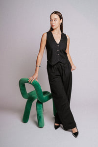 Woman in black sleeveless top and pants standing next to a green chair on a plain background