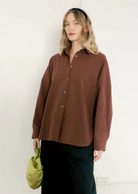 Woman wearing wine stripe oversized shirt, black pants, and holding green bag against white wall