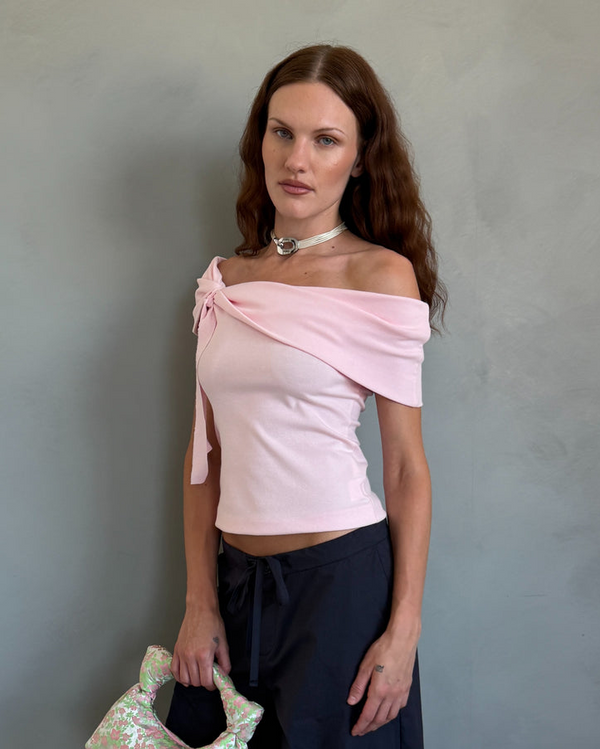 Woman wearing a pink off-shoulder top and dark pants, holding a floral clutch against a gray wall.