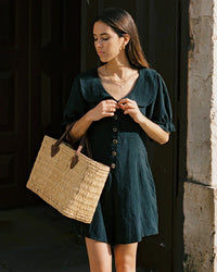 Woman in black linen romper with buttons, holding woven straw tote bag outdoors