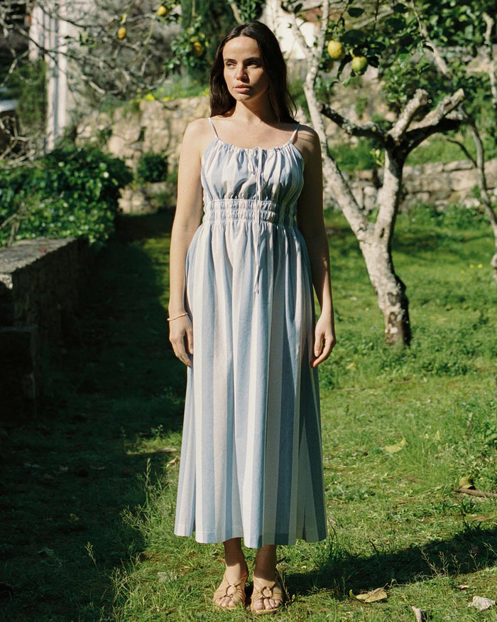 Woman in a light blue dress standing in a garden with trees and stone wall in the background