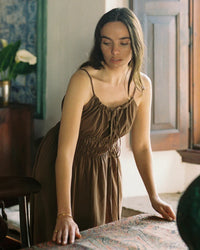 Woman in mocha cotton Sicily dress stands indoors by sunlit window and table